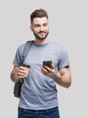 Sırt çantasında akıllı telefon ve gri arka planda izole kahve tutan yakışıklı bir delikanlı. Ciddi öğrenci ya da iş adamı portresi. Neşeli adamlar seyahate çıkıyor.
