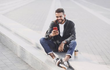 Şehirde akıllı telefon kullanan genç yakışıklı adamlar. Gülümseyen öğrenci cep telefonuyla mesaj atıyor. Kahve molası. Modern yaşam tarzı, bağlantı, iş konsepti