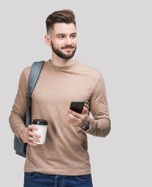 Sırt çantasında akıllı telefon ve gri arka planda izole kahve tutan yakışıklı bir delikanlı. Ciddi öğrenci ya da iş adamı portresi. Neşeli adamlar seyahate çıkıyor.