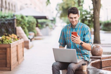 Genç yakışıklı iş adamı şehirde dizüstü bilgisayar ve akıllı telefon kullanıyor. Gülümseyen öğrenci kahve molası veriyor. Modern yaşam tarzı, bağlantı, iş konsepti