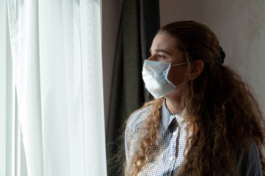 Tıbbi maskeli ve eldivenli genç bir kız kendini tecrit ederken pencereden dışarı bakar, evde karantinaya alınır. Salgın, grip salgını, viral hastalıklar, Covid-19..