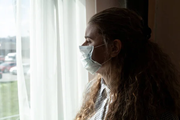 Tıbbi maskeli ve eldivenli genç bir kız kendini tecrit ederken pencereden dışarı bakar, evde karantinaya alınır. Salgın, grip salgını, viral hastalıklar, Covid-19..