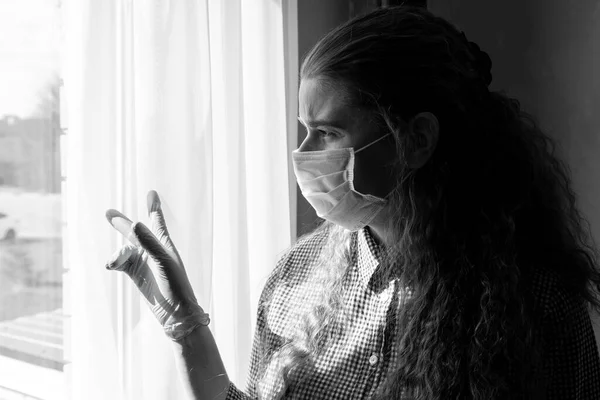 Tıbbi maskeli ve eldivenli genç bir kız kendini tecrit ederken pencereden dışarı bakar, evde karantinaya alınır. Salgın, grip salgını, viral hastalıklar, Covid-19..