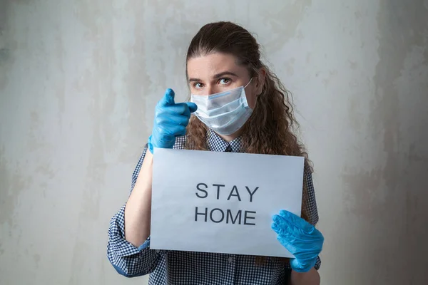 A young girl in a medical mask and gloves with an inscription on a sheet of paper calls for staying in quarantine at home. Home self-isolation in pandemic, flu epidemic, viral diseases, Covid-19.