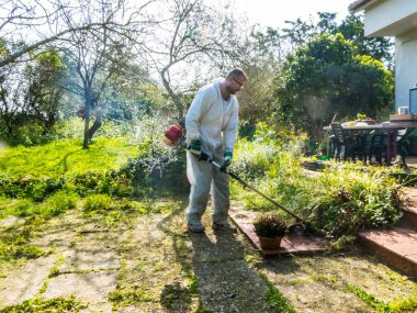 Beyaz takım elbiseli bir adam güneşli bir günde bahçedeki çimleri biçiyor.
