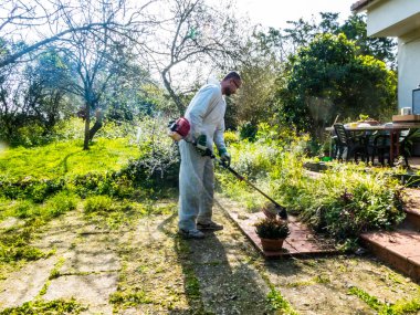 Beyaz takım elbiseli bir adam güneşli bir günde bahçedeki çimleri biçiyor.