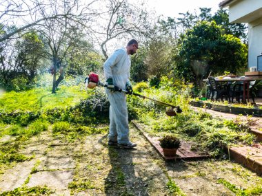 Beyaz takım elbiseli bir adam güneşli bir günde bahçedeki çimleri biçiyor.
