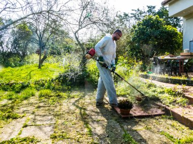 Beyaz takım elbiseli bir adam güneşli bir günde bahçedeki çimleri biçiyor.