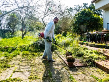 Beyaz takım elbiseli bir adam güneşli bir günde bahçedeki çimleri biçiyor.
