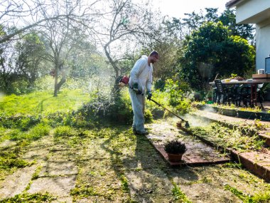 Beyaz takım elbiseli bir adam güneşli bir günde bahçedeki çimleri biçiyor.