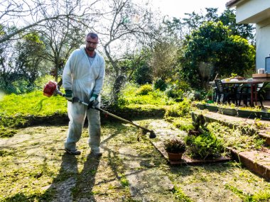 Beyaz takım elbiseli bir adam güneşli bir günde bahçedeki çimleri biçiyor.