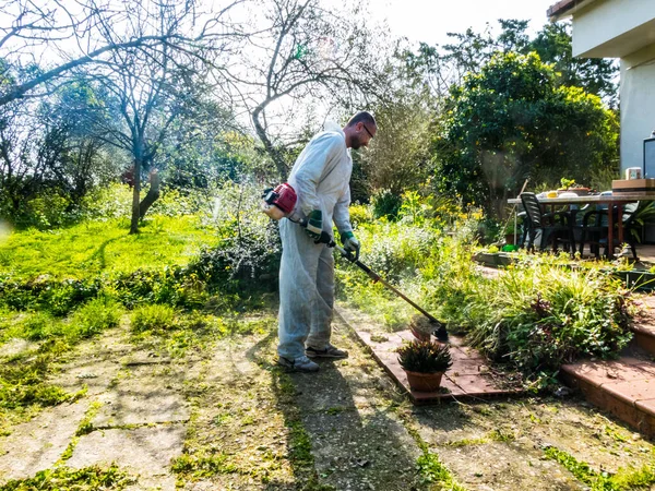 Beyaz takım elbiseli bir adam güneşli bir günde bahçedeki çimleri biçiyor.