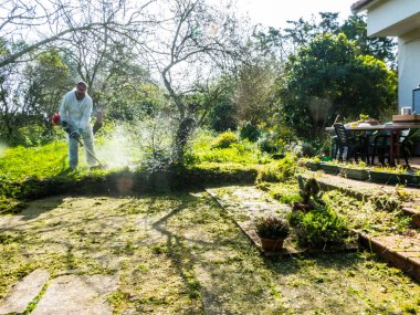 Beyaz takım elbiseli bir adam güneşli bir günde bahçedeki çimleri biçiyor.