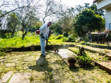 Beyaz takım elbiseli bir adam güneşli bir günde bahçedeki çimleri biçiyor.