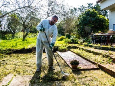 Beyaz takım elbiseli bir adam güneşli bir günde bahçede demir süpürgeyle temizlik yapıyor.