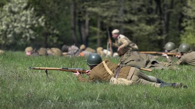 Miğfer ve tüfekle yere saldıran asker, Rus piyadelerine cepheden saldıran, ikinci dünya savaşını yeniden inşa eden. Arka planda daha fazla asker bulanıklaşıyor. Çayır ve orman.