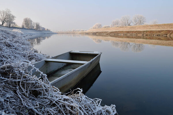 Abandoned row boat moored by the river bank in winter. Calm river, morning, sun and frost.