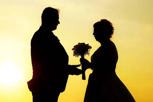 Silhouette of groom and bride and wedding bouquet between them, orange sunlight background behind them. Hair and flower shines through the golden light.
