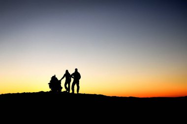 Norveç 'te gece yarısı gün batımı, taş sütunu, koyu mavi ve turuncu alacakaranlık gökyüzü, siyah ön plan ve doymuş gökyüzü olan kadın ve erkek silueti. Dağlarda alacakaranlık, açık gökyüzü.