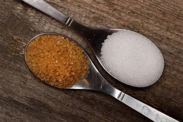 Two spoons with cane sugar and white sugar next to each other on wooden board. Comparison of sugars.