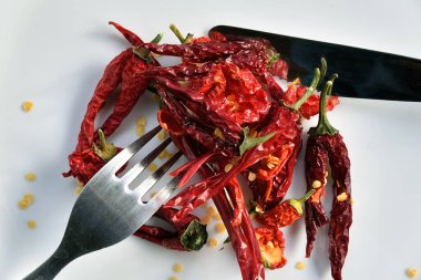 Kurutulmuş kırmızı biberler beyaz tabakta, tohumlar etrafa saçılmış, çatal ve bıçak biberde.