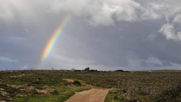 Malta island and rainbow after rain near of golden bay, country roat between bush and stones. Rain and sun, Land near ghajn tuffieha. Dark stormy blue sky, sunlight and part of rainbow.