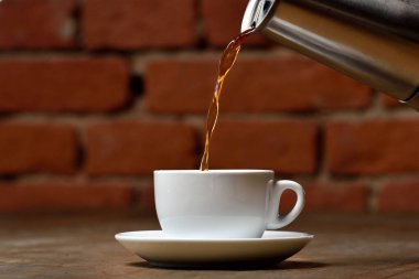 Kahve makinesinin çay tabağıyla beyaz porselen bardağa kahve dolduruyorum. Kupa ahşap masada ve paslanmaz çaydanlıktan gelen kahve akıntısı. Ahşap masa ve tuğla duvar arkaplanı.