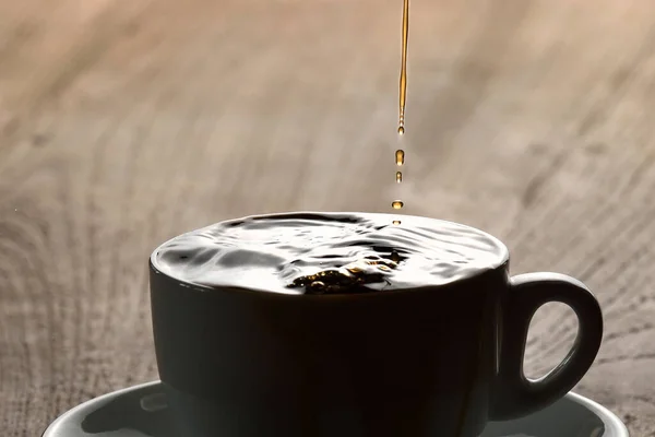 Pouring coffee into white porcelain mug with saucer from moka pot coffee maker. Mug on wooden table and stream of coffee from kettle. Stormy shiny black surface of full coffee mug and coffee drops.