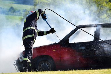 Araba yangını. Bir itfaiyeci araba yangınını köpükle söndürür. Kırmızı araba yanıyor, beyaz duman ve itfaiyeci. Çayırda bir kaza oldu..