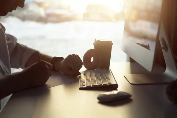 The guy working at the computer and drinking coffee with a donut