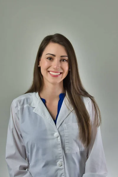 Gri duvarda izole edilmiş beyaz önlüklü kadın sağlık uzmanı. Kafa ve omuzlar görünür. Tarafsız zemin üzerinde güzel gülüşü olan çekici genç bayan.