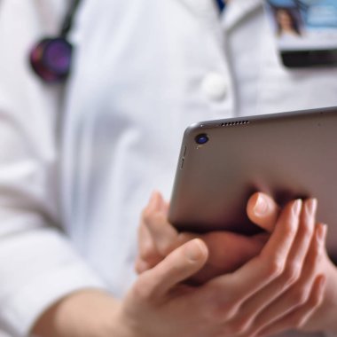 Tablet, yakından sağlık uzmanının elinde. Beyaz önlük, steteskop ve arka planda görünen bir rozet. Pratisyen hemşirelerin ya da PA 'nın elleri, tıp teknolojisini hastalar için kullanıyor.