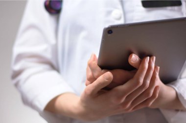 Tablet, yakından sağlık uzmanının elinde. Beyaz önlük, steteskop ve arka planda görünen bir rozet. Pratisyen hemşirelerin ya da PA 'nın elleri, tıp teknolojisini hastalar için kullanıyor.