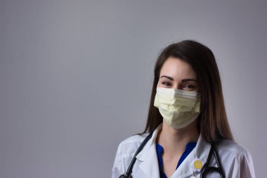 Masked woman medical professional smiling under mask. Isolated on grey background with white coat, stethoscope, and yellow PPE mask visible clipart