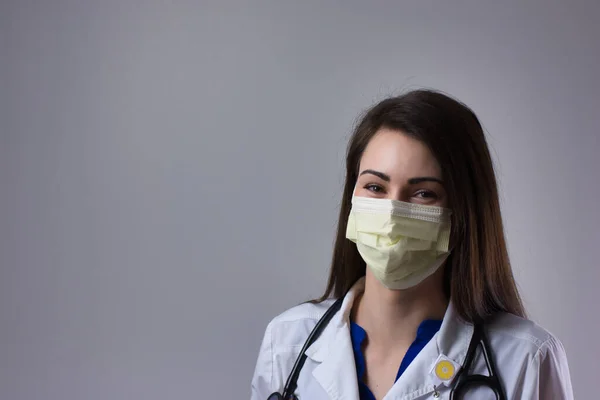 Maske altında gülümseyen maskeli kadın tıp uzmanı. Beyaz önlük, steteskop ve sarı PPE maskeli gri arka planda izole edilmiş.