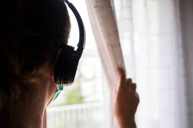 Kız pencereden dışarı bakıyor ve koruyucu maske ve eldivenlerle korunan müzik dinliyor. Coronavirus, Covid-19