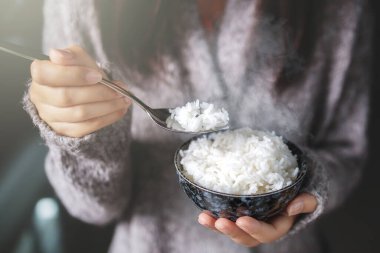 Asyalı kadınlar ellerinde kaşık ile pişirilmiş yasemin kaseleri tutuyorlar. Gri bir kazak giyiyor..