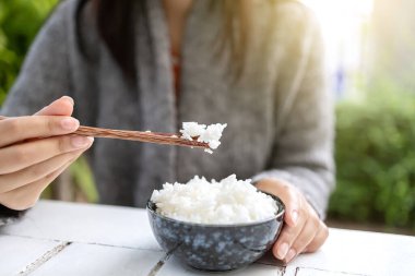 Asyalı kadınlar ellerinde yemek çubuklarıyla pişirilmiş yasemin kaseleri tutuyorlar. Gri bir kazak giyiyor..