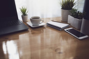 Sabahları ofis penceresinden güneş ışığı alıyor. Laptop, beyaz kahve fincanı, defter ve çalışan masadaki akıllı telefonla..