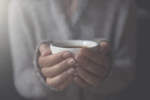 Closeup of female hands with a cup of hot coffee. in the morning soft sunlight. Beautiful girl in grey sweater.