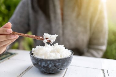Asyalı kadınlar ellerinde yemek çubuklarıyla pişirilmiş yasemin kaseleri tutuyorlar. Gri bir kazak giyiyor..