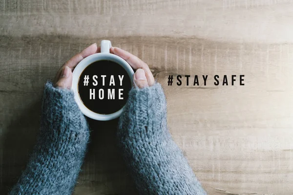 Woman\'s hands in gray sweater holding cup of black coffee with text hashtag #STAY HOME and #STAY SAFE on the wooden table. Concept stay home stop virus.