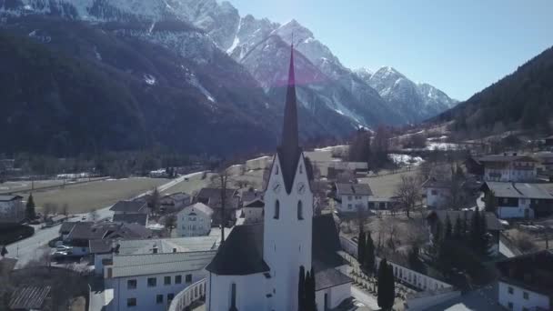 Widok z lotu ptaka na kościół w małym miasteczku pomiędzy potężnymi górami w Alpach — Wideo stockowe