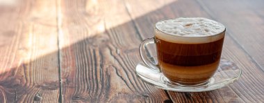 Kapuçino, şeffaf bir bardakta, güneş ışığında, tahta bir masanın üzerinde.