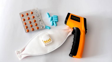 Beyaz arka planda, hapların ve solunum maskesinin yanında elektronik termometre var..