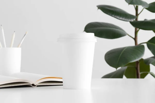 Coffee paper cup mockup with a notebook, workspace accessories and a ficus plant on a white table.