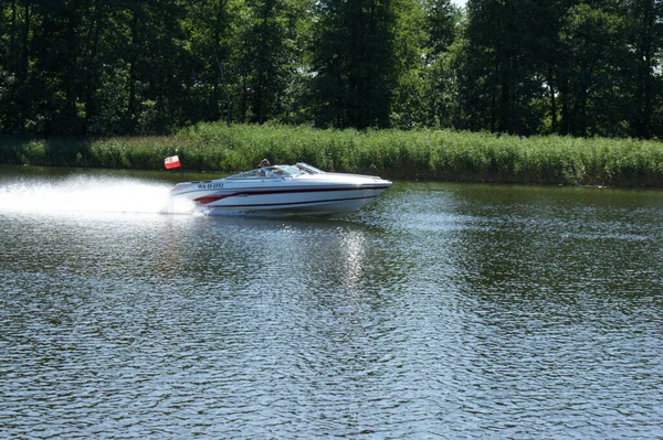 白色的汽艇沿着绿岸的草地和树林在湖面上疾驰而过 免版税图库照片