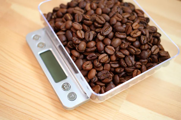 Weighing beautiful fresh dark roasted coffee beans in plastic transparent container on electronic digital silver kitchen scale with liquid crystal screen, against light brown wooden surface