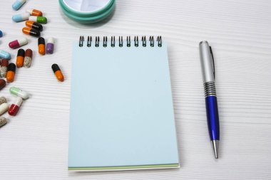 Bir grup çok renkli ilaç kapsülü yeşil bir hap kutusu ve boş mavi bir not defterinin yanında.