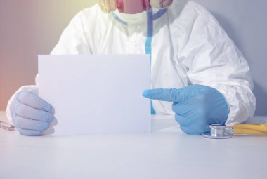 Solunum cihazı ve eldiven takmış biyolojik tehlike kıyafeti giymiş bir doktor boş bir kağıt parçasını işaret ediyor.